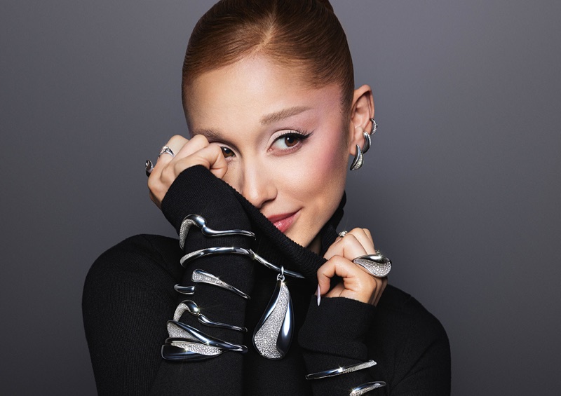 Ariana Grande in a black turtleneck sweater wearing sculptural Swarovski silver jewelry against a grey studio backdrop.