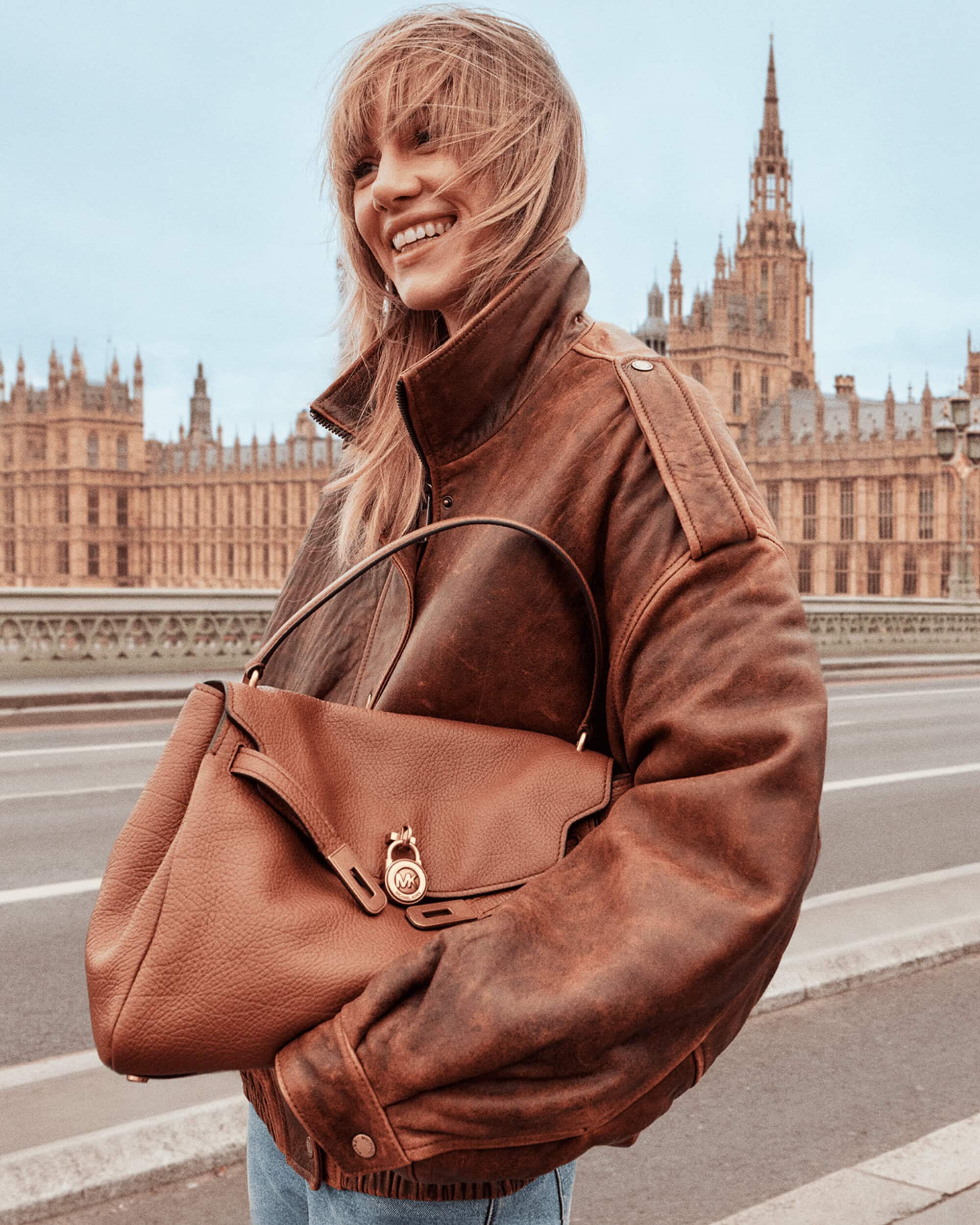 Smiling woman in an oversized distressed brown leather jacket holding a brown shoulder bag with historic architecture in the background.