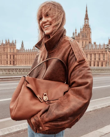 Smiling woman in an oversized distressed brown leather jacket holding a brown shoulder bag with historic architecture in the background.