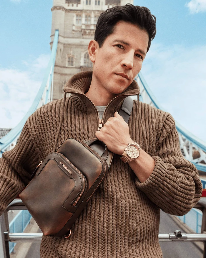 Man in a brown ribbed sweater wearing a leather sling bag and gold watch on a bridge in London.