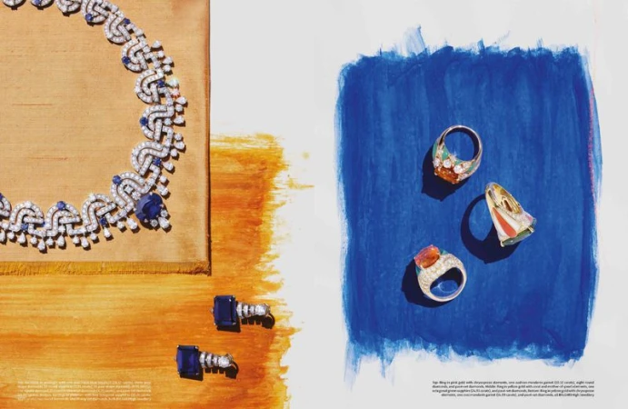 High-jewelry product still-life: a diamond and sapphire necklace and stud earrings displayed on a gold textured surface, and three diamond rings on blue backdrop.