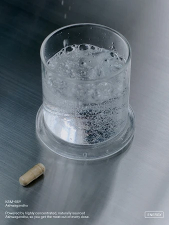 Glass of bubbling water beside an ashwagandha supplement capsule on a brushed metal surface.