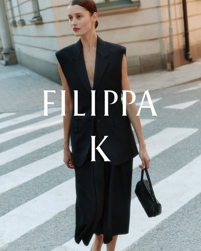 Model wearing a black sleeveless blazer and trousers holding a handbag on a city street.