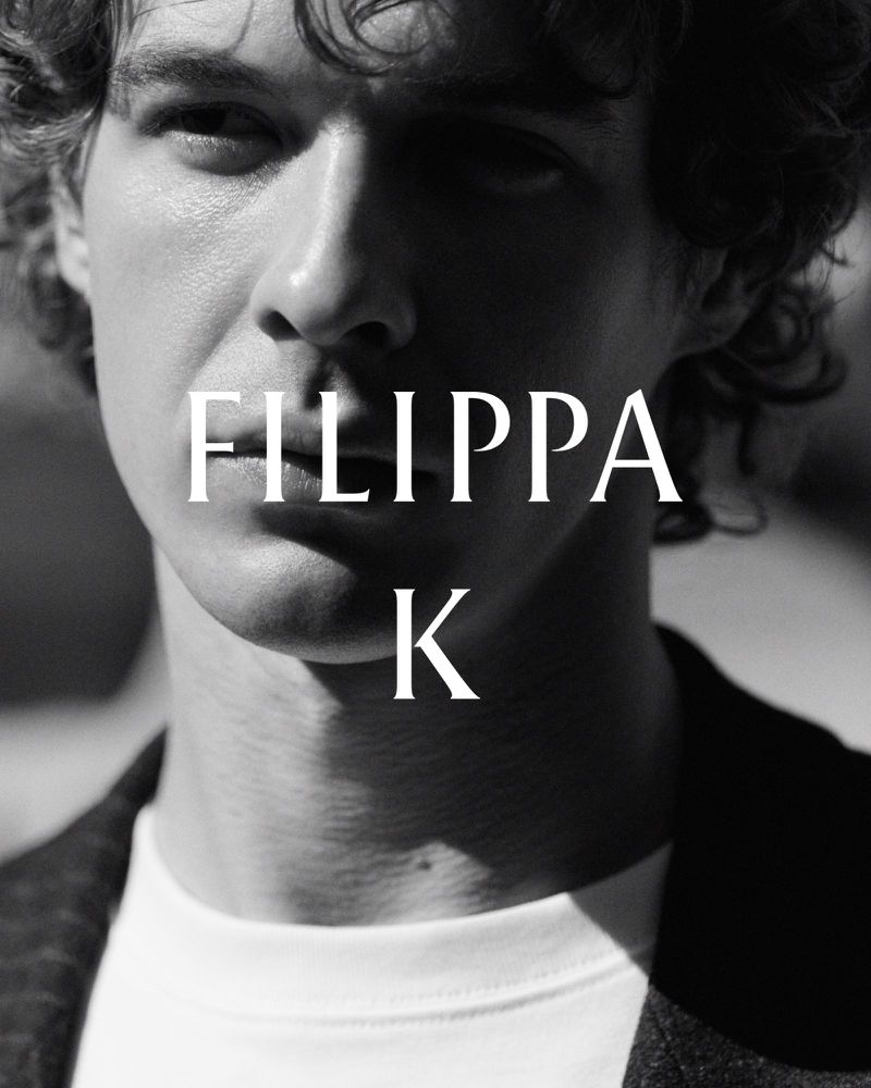 Close-up black and white portrait of a man with curly hair wearing a white t-shirt and dark jacket with Filippa K text overlay.