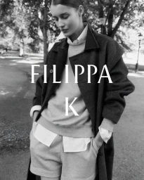 Model wearing a layered collared shirt, crewneck sweater, shorts, and an oversized coat in black and white for Filippa K.