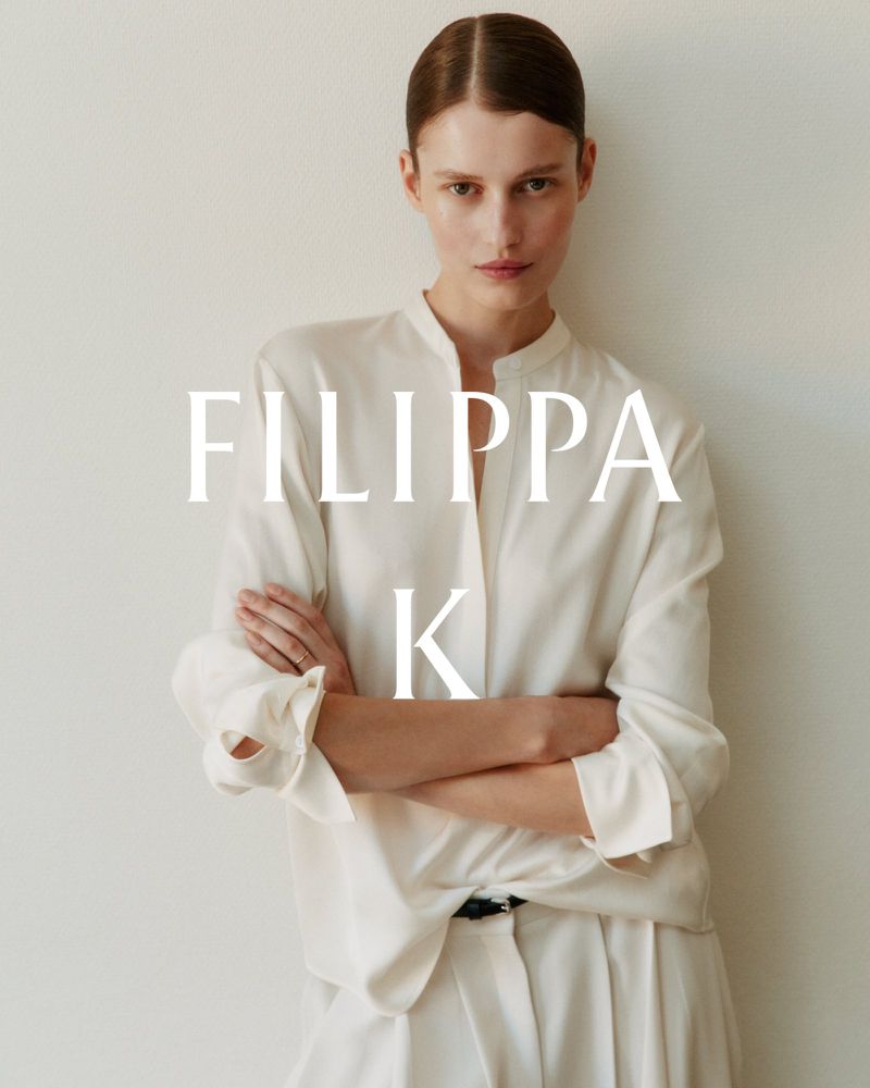 Model with slicked back hair posing with arms crossed in an off-white silk blouse and trousers against a white studio backdrop.