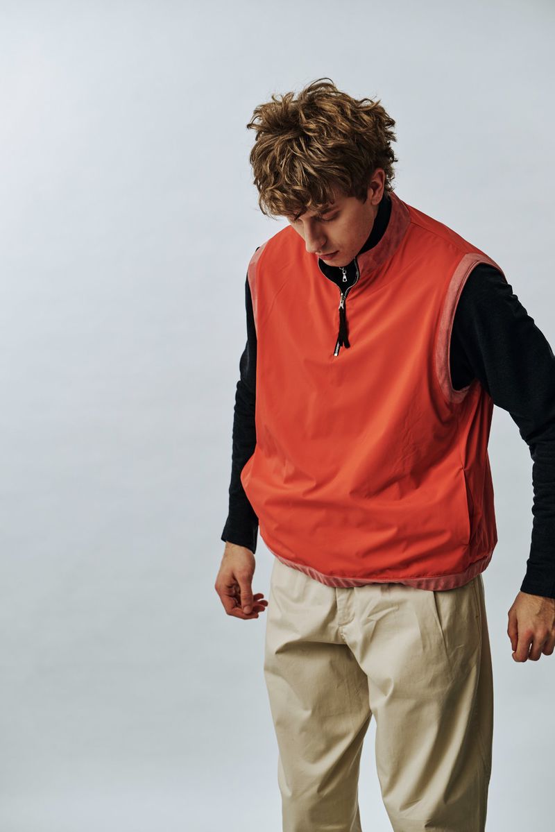 Model wearing an orange zip vest over a black long-sleeve top with beige trousers against a pale studio backdrop.