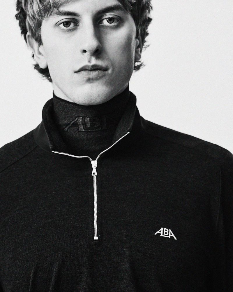 Monochrome studio portrait of a man wearing a black ALBA half-zip pullover and high-neck top.