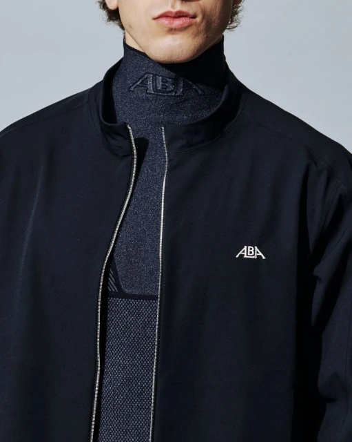 Cropped shot of a man wearing a dark zip-up jacket over a high-neck textured base layer with an ABA logo against a neutral background.