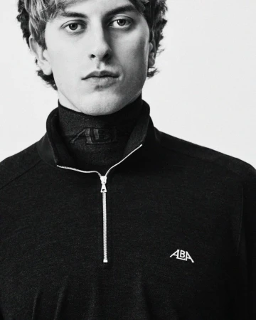 Close-up portrait of a young man wearing a black ALBA quarter-zip pullover over a turtleneck against a white backdrop.