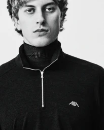 Close-up portrait of a young man wearing a black ALBA quarter-zip pullover over a turtleneck against a white backdrop.