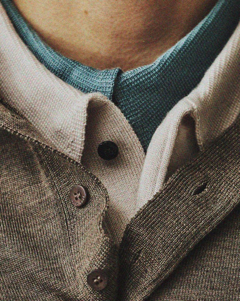 Close-up of layered menswear knitwear featuring a taupe buttoned cardigan over a beige polo and teal inner collar.