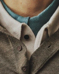 Close-up of layered menswear knitwear featuring a taupe buttoned cardigan over a beige polo and teal inner collar.