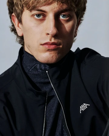 Close-up portrait of a man with blue eyes wearing a black zip jacket with a white logo against a pale studio backdrop.