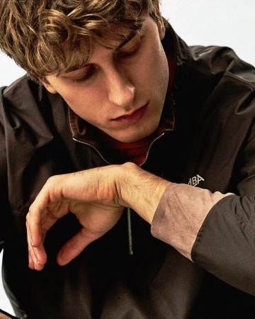 Young man looking down while wearing a dark zip-up jacket with a taupe cuff against a white studio background.