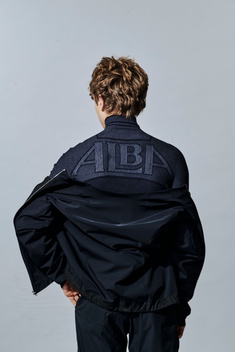 Model seen from behind wearing an ALBA athletic turtleneck top and dark windbreaker jacket against a neutral studio background.
