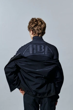 Model seen from behind wearing an ALBA athletic turtleneck top and dark windbreaker jacket against a neutral studio background.