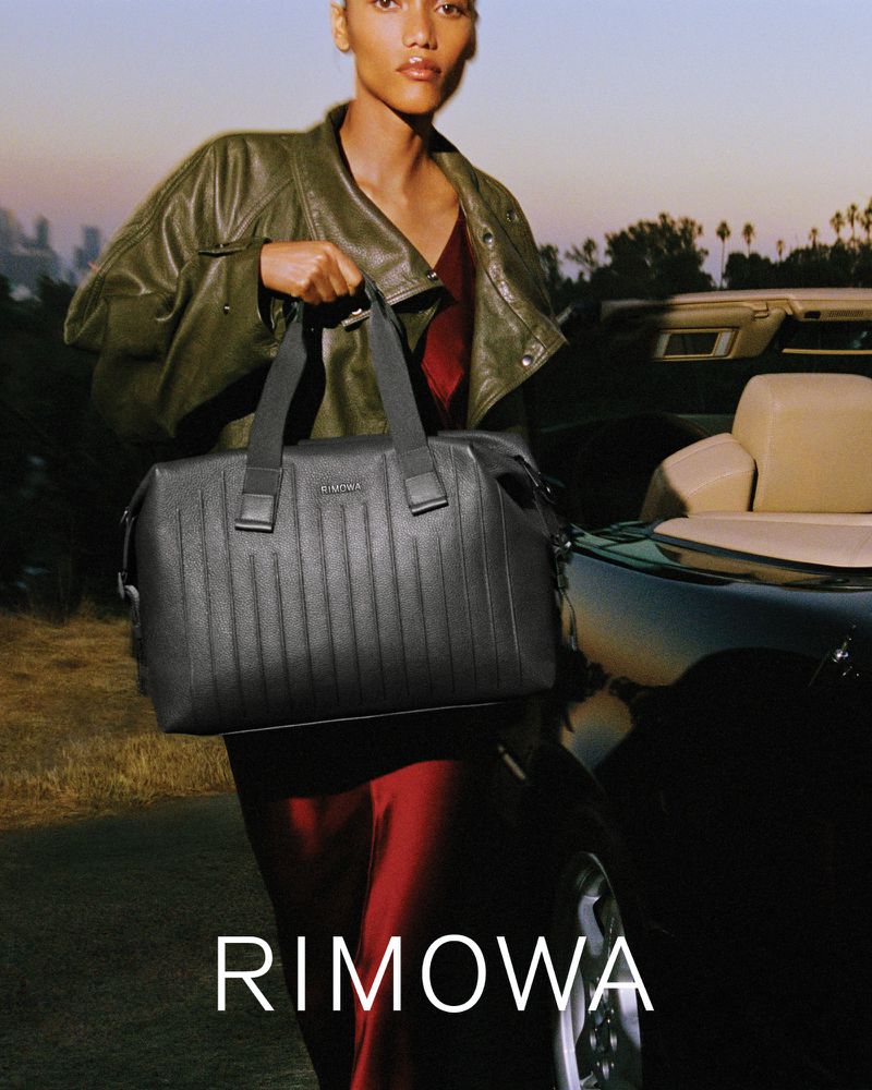 Model carrying a black grooved leather duffle bag while standing beside a dark convertible car at dusk.