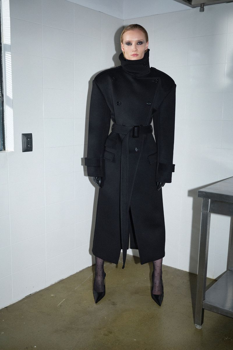 Model posing in an oversized black belted high-collar trench coat with pointed heels in a tiled interior corner.