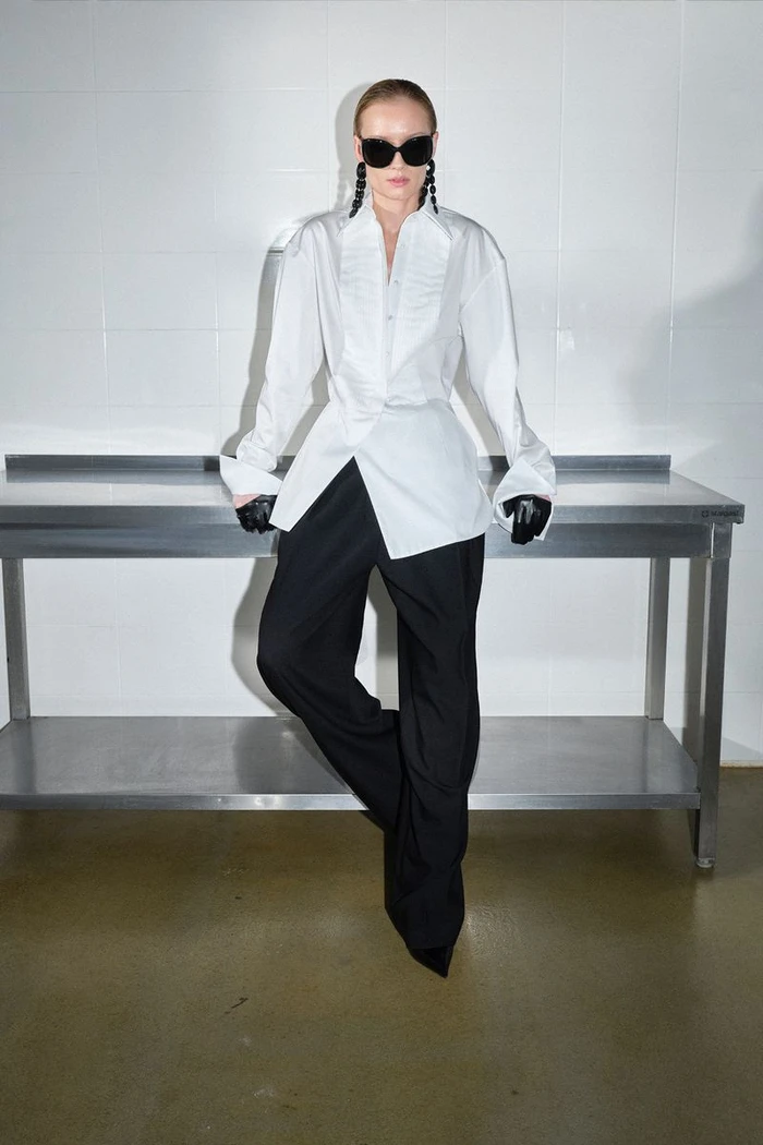 Model posing in a structured white button-down shirt, black wide-leg trousers, sunglasses, and gloves against a steel prep table.