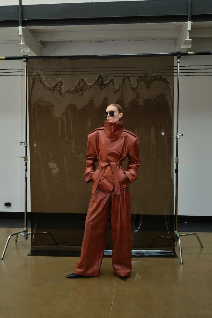 Model Kasia Smulska wearing a terracotta leather belted jacket and wide-leg trousers with sunglasses in front of a studio backdrop.