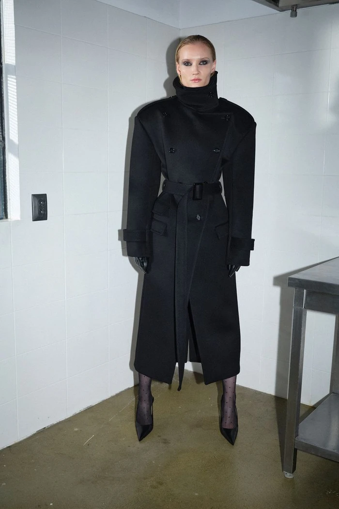 Model Kasia Smulska standing in a corner wearing an oversized black belted coat, black gloves, and pointed black heels.