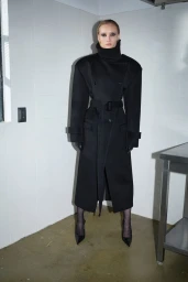 Model Kasia Smulska standing in a corner wearing an oversized black belted coat, black gloves, and pointed black heels.