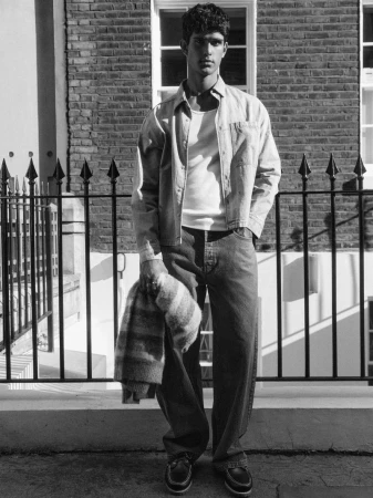 Model Earl Cordeiro posing in an open button-up shirt and relaxed jeans against a brick building and iron fence.