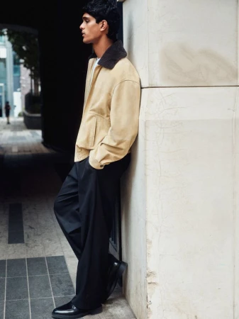 Model Earl Cordeiro leaning against a stone pillar wearing a beige shearling collar jacket and wide leg black trousers.