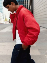 Model Earl Cordeiro walking along a city sidewalk wearing a vibrant red zip-up bomber jacket, white shirt, and dark denim jeans.