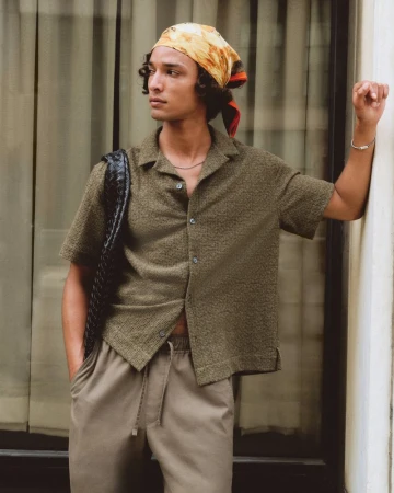 Model Djairo Mulder wearing an olive textured short-sleeve shirt, headscarf, and taupe trousers leaning against a white pillar.