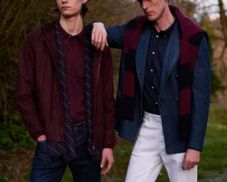 Two male models posing outdoors in layered menswear including a burgundy jacket, denim blazer, and white trousers.