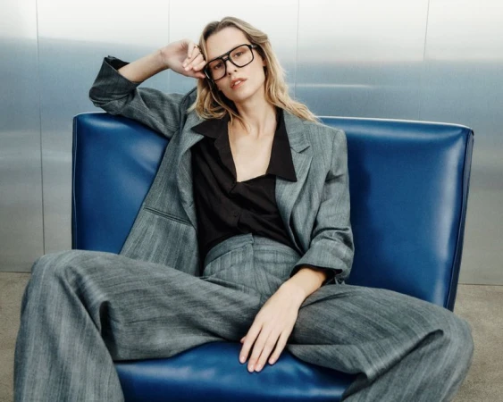 Model Mariina Keskitalo in a grey pinstripe suit and black eyeglasses seated in a vibrant blue armchair against a brushed metal wall.