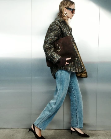 Model Mariina Keskitalo walking in a tweed jacket, blue jeans, black kitten heels, and holding a brown suede bag against a metal wall.