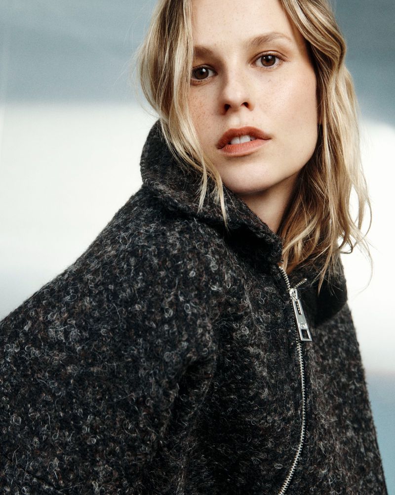 Mariina Keskitalo wearing a heavy textured charcoal zip-up coat looking over her shoulder against a soft light background.