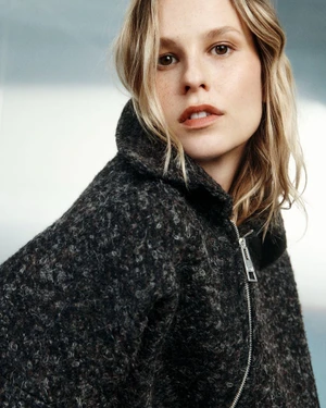 Mariina Keskitalo wearing a heavy textured charcoal zip-up coat looking over her shoulder against a soft light background.