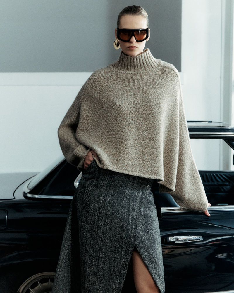 Model Mariina Keskitalo wearing an oversized taupe turtleneck poncho and slit skirt beside a black vintage car.