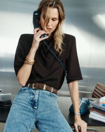Model Mariina Keskitalo talks on a retro landline phone while sitting on a desk in high-waisted jeans and a shoulder-padded top.