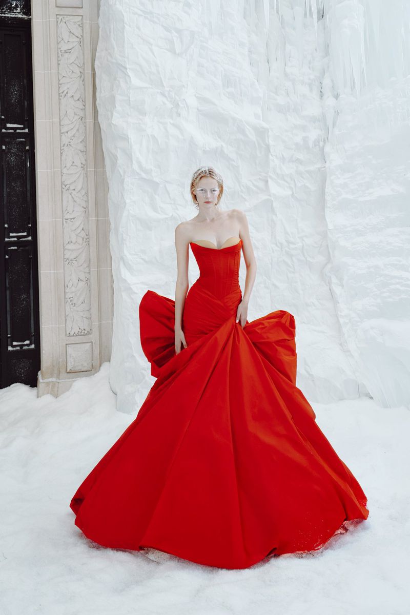 Model Elza Matiz wearing a voluminous red strapless couture ballgown standing in snow against a sculpted ice wall.