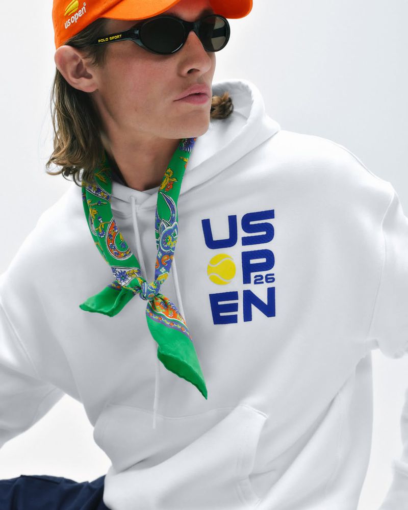 Male model wearing an orange cap, black sunglasses, a green neckerchief, and a white US Open graphic hoodie in studio.