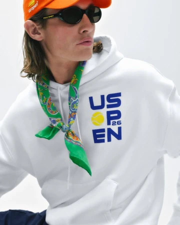 Male model wearing an orange cap, black sunglasses, a green neckerchief, and a white US Open graphic hoodie in studio.