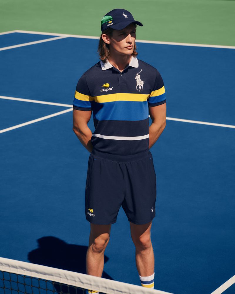 A man wearing a navy Polo Ralph Lauren US Open polo shirt, athletic shorts, and a matching cap stands on a blue tennis court.