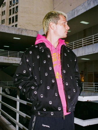 Blonde man wearing a black printed zip-up jacket over a bright pink hoodie standing near a white balcony railing.