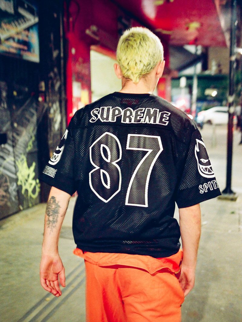 Rear view of a man with dyed blonde hair wearing a black Supreme Spitfire 87 mesh jersey and orange pants on a city sidewalk.