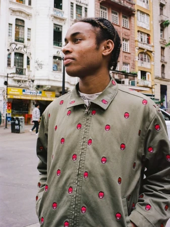 Young Black man with braided hair looking away outdoors while wearing an olive zip jacket with red graphic pattern.