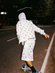 Skater riding a skateboard down an asphalt road at night wearing a white printed Supreme hooded jacket and matching shorts.