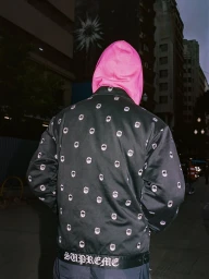 Person seen from behind wearing a vibrant pink hoodie and a black printed Supreme Spitfire jacket on an urban street at night.