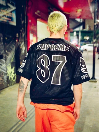 Man with bleached hair seen from behind wearing a black Supreme Spitfire 87 mesh jersey and orange pants on a city street.