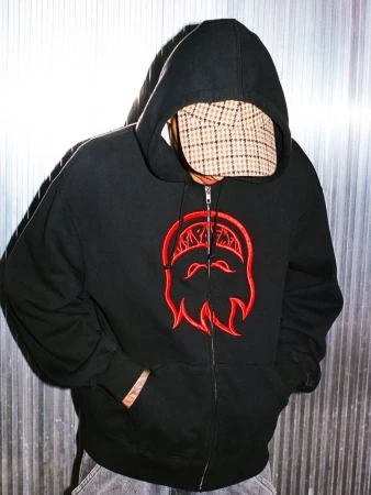 Model wearing a black zip-up hooded sweatshirt with red embroidered graphic and plaid cap in front of corrugated metal.