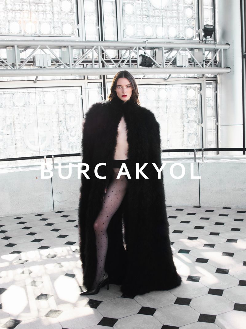 Ansley Gulielmi wearing an oversized black fur cape and sheer tights against a geometric tiled floor and glass wall.